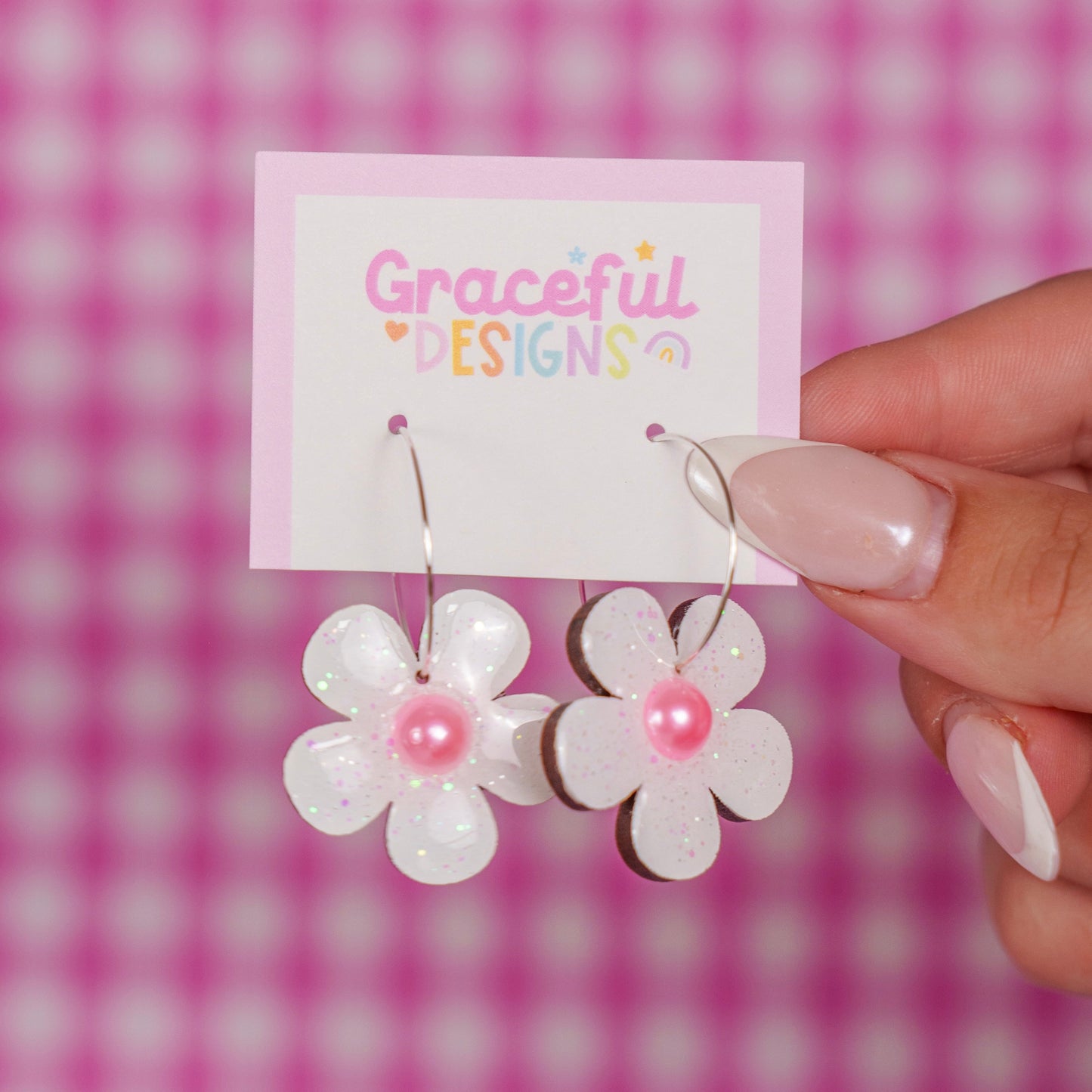 Flower-shaped earrings with pink centers held by a hand against a pink checkered background.
