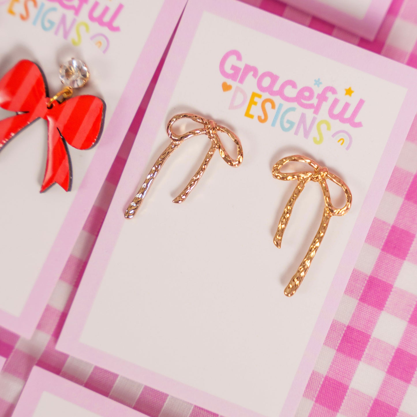 Gold bow-shaped earrings on a 'Graceful Designs' branded card with a pink checkered background.