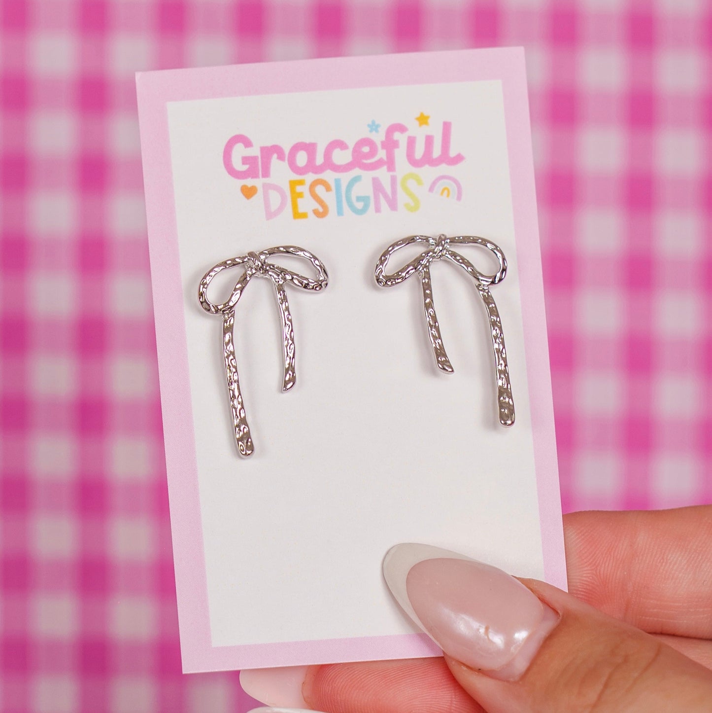 Silver bow-shaped earrings on a 'Graceful Designs' card against a pink checkered background.