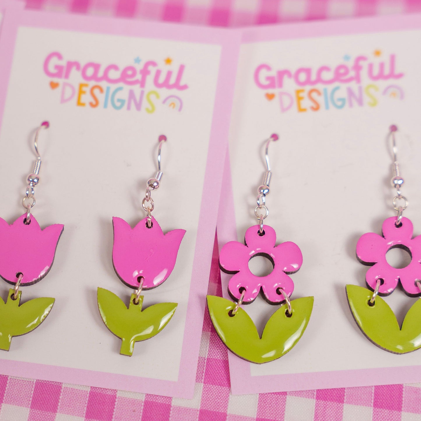 Pair of pink and green floral earrings on 'Graceful Designs' cards against a pink checkered background.