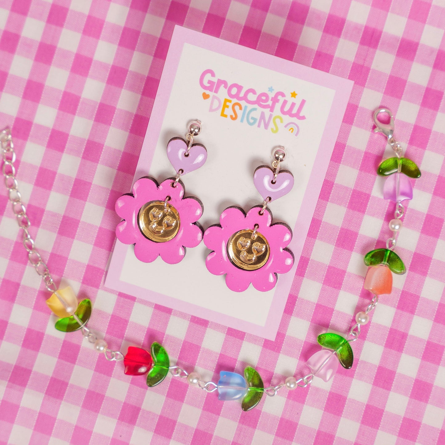 Jewelry set with pink flower-shaped earrings and a matching necklace on a 'Graceful Designs' card against a pink checkered background.