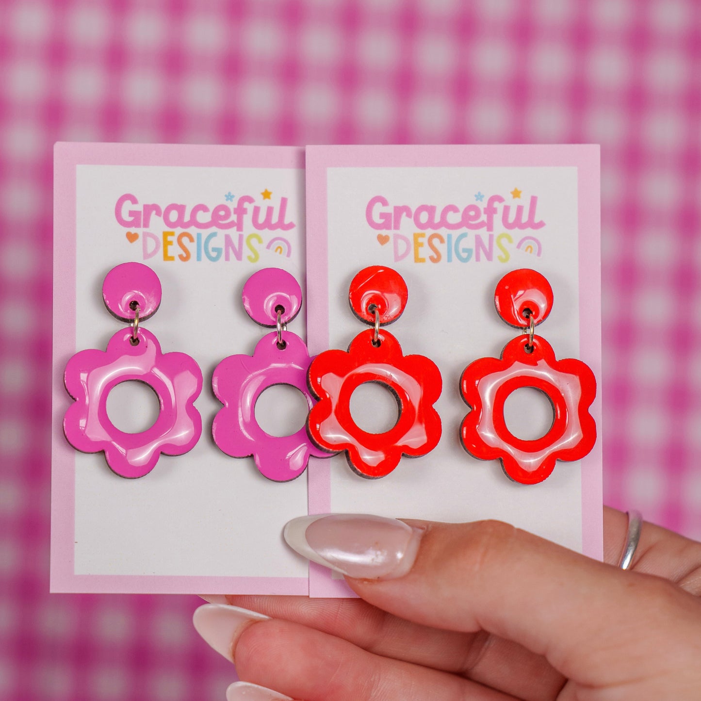 Pair of pink and red floral earrings on 'Graceful Designs' cards held by a hand against a pink checkered background.