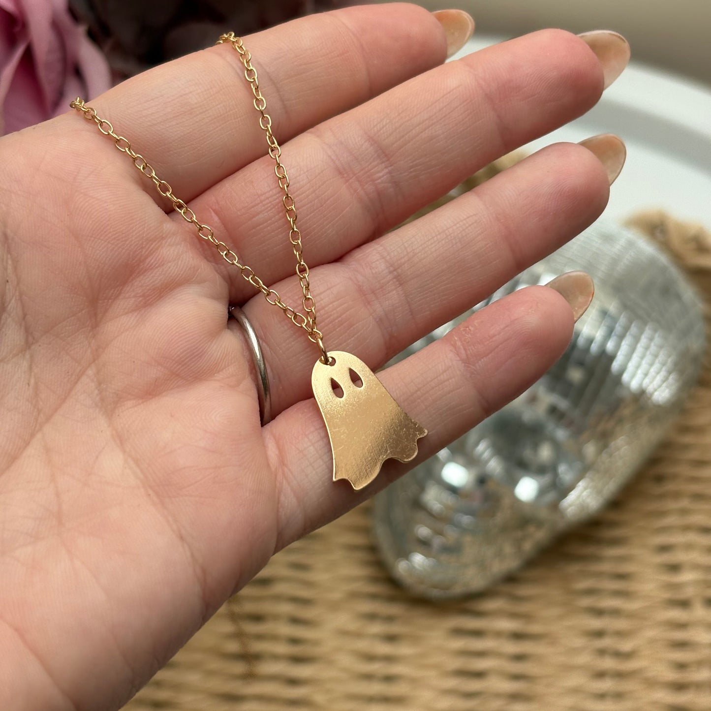 Hand wearing a gold necklace with a ghost-shaped pendant against a neutral background