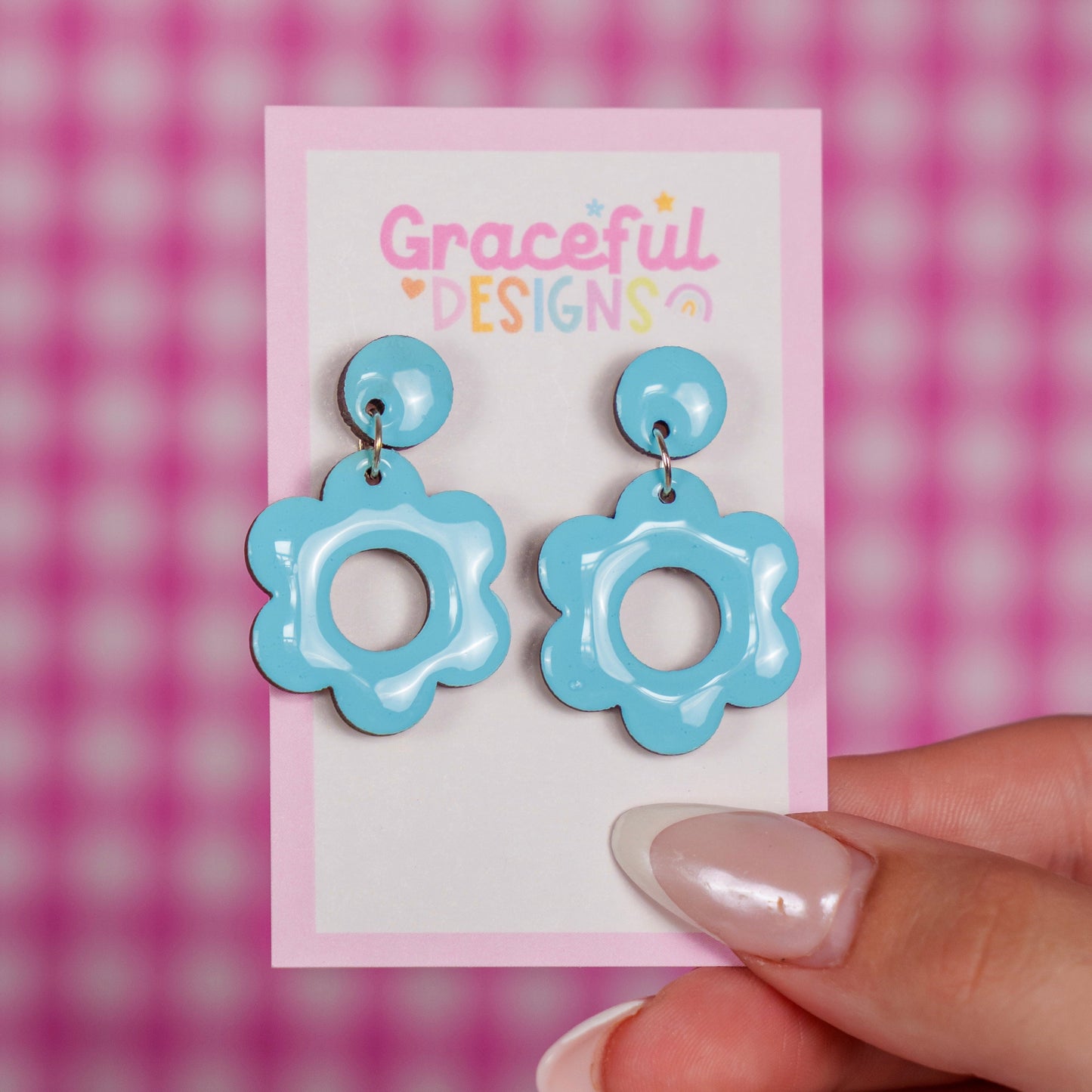 Blue flower-shaped earrings on a card with 'Graceful Designs' branding, held against a pink checkered background.