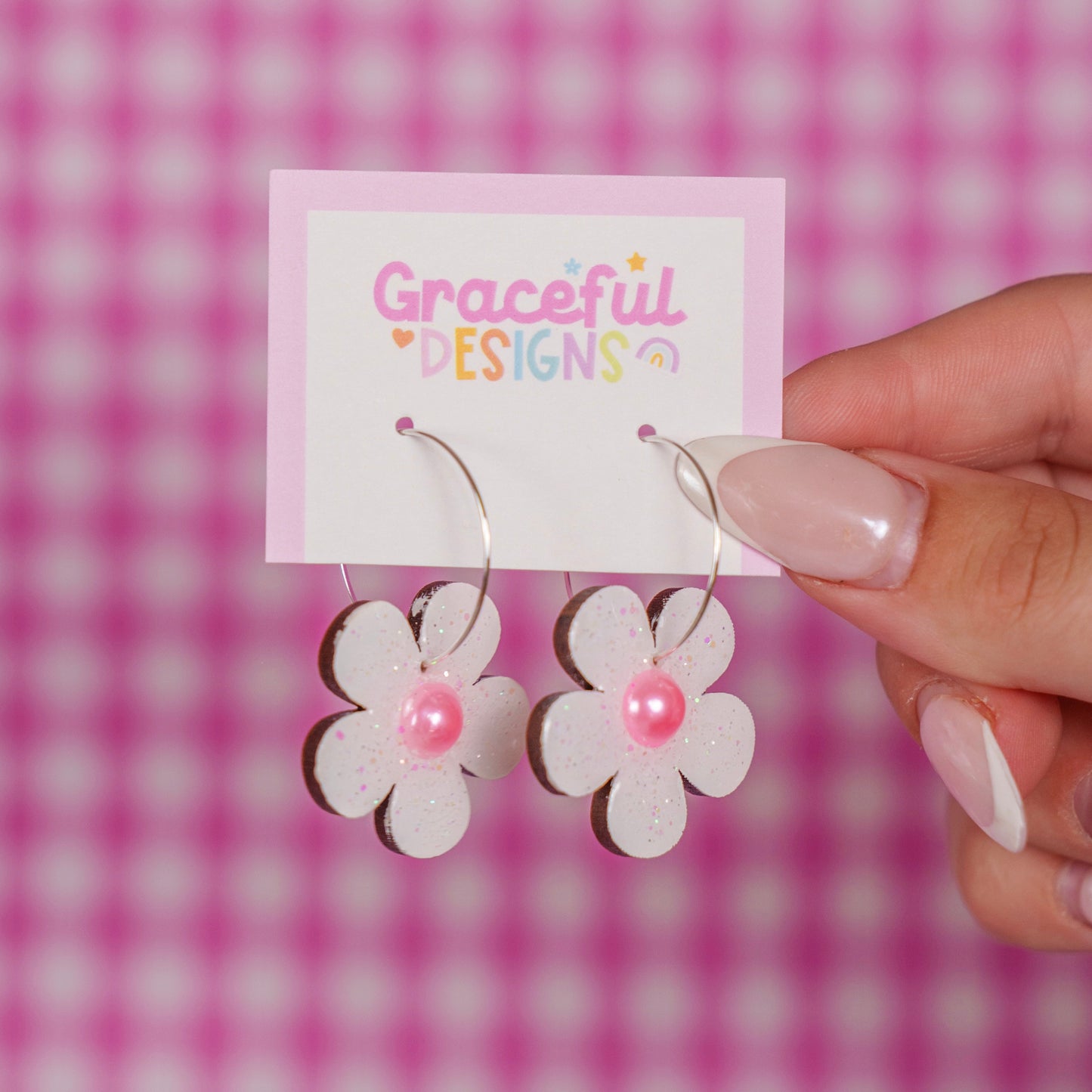 Flower-shaped earrings on a 'Graceful Designs' card against a pink checkered background
