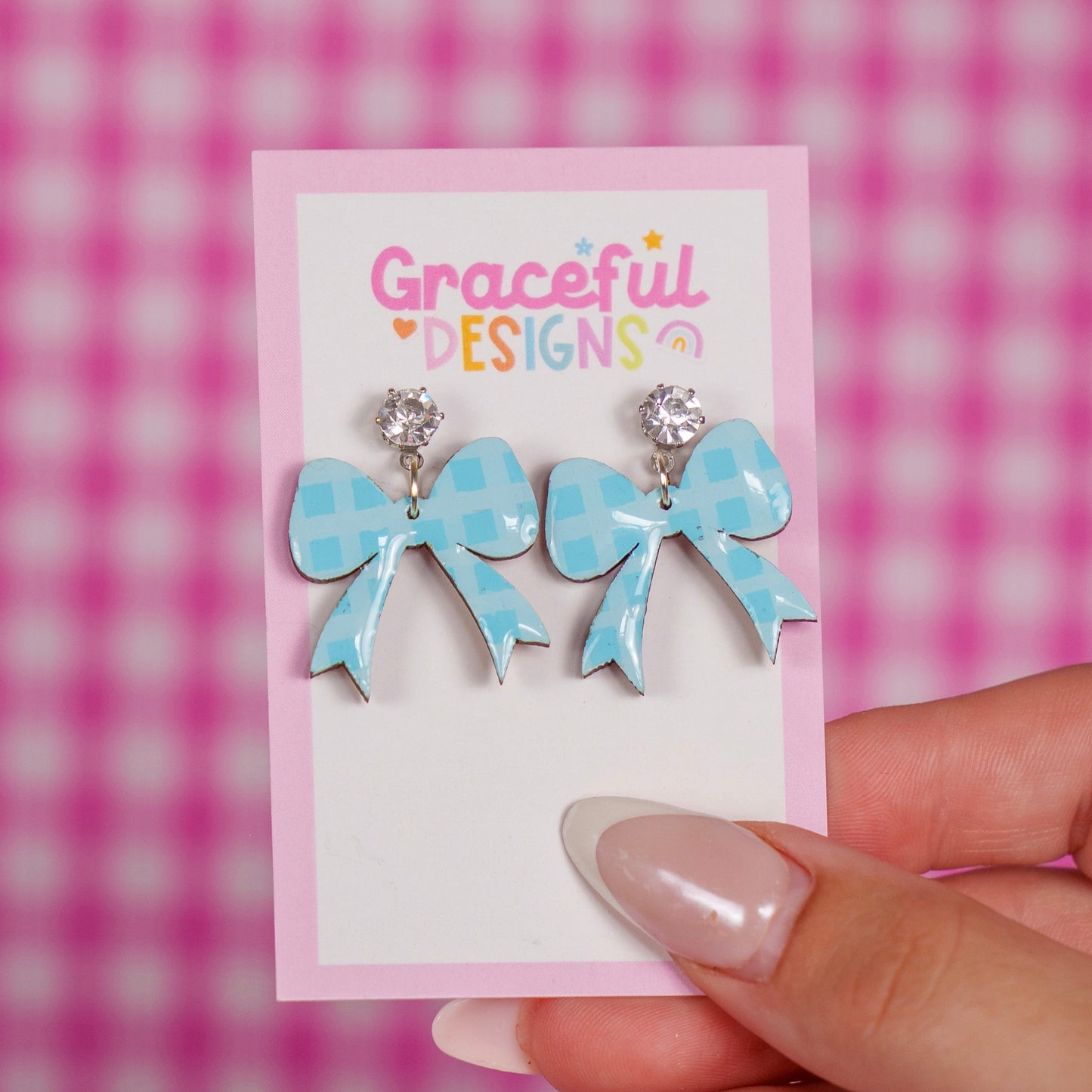Blue bow-shaped earrings on a 'Graceful Designs' card against a pink checkered background.