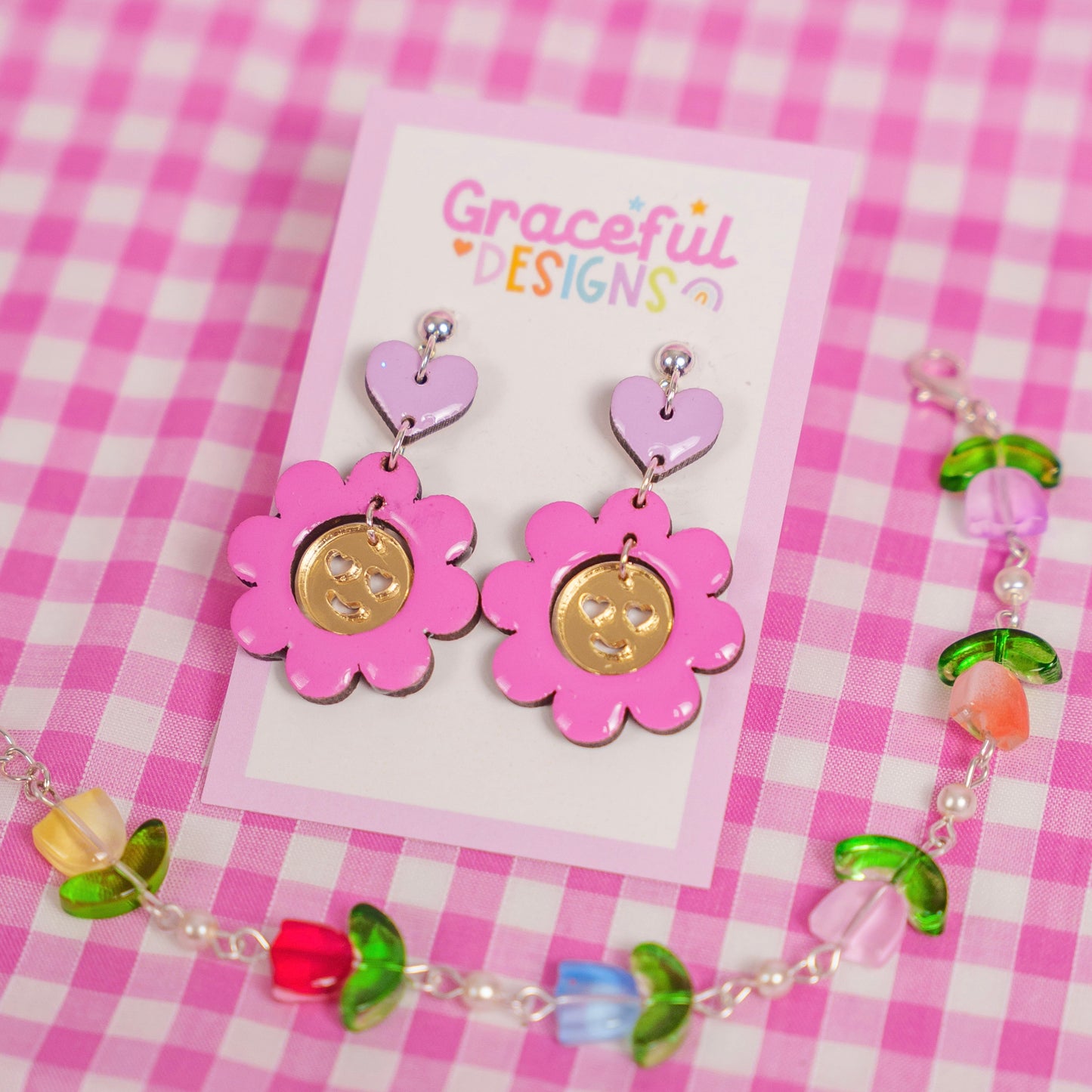 Pair of pink flower-shaped earrings on a 'Graceful Designs' card against a pink checkered background.
