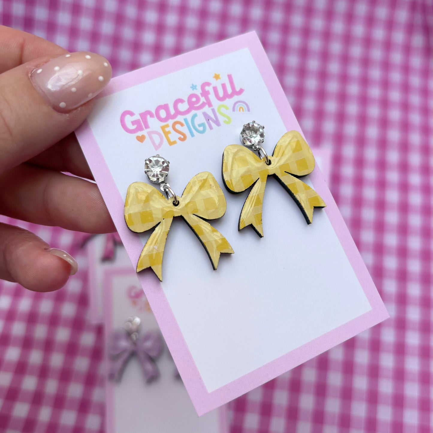 Yellow bow-shaped earrings on a 'Graceful Designs' card against a pink checkered background.