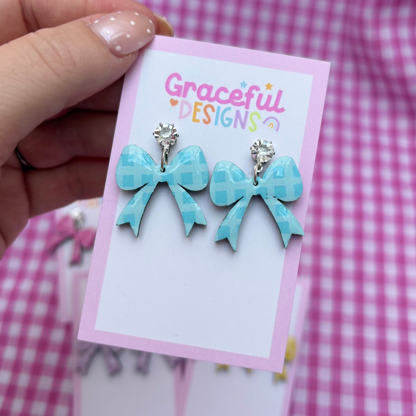 Blue bow-shaped earrings on a 'Graceful Designs' card against a pink checkered background