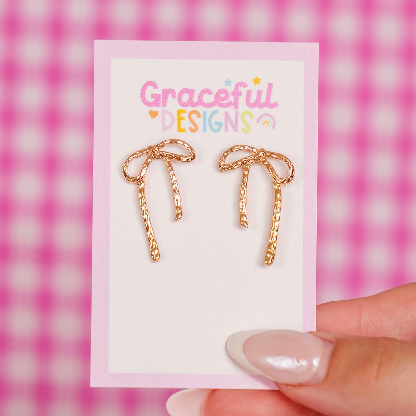 Gold bow-shaped earrings on a 'Graceful Designs' card against a pink checkered background.
