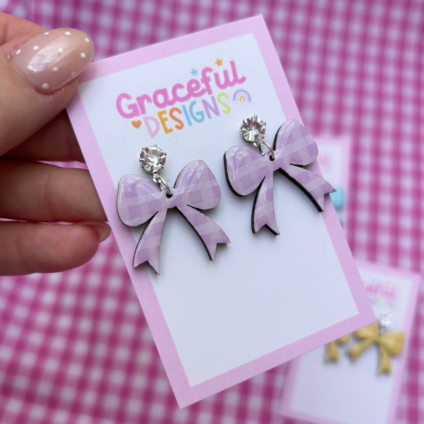 Purple bow earrings on a 'Graceful Designs' card against a pink checkered background