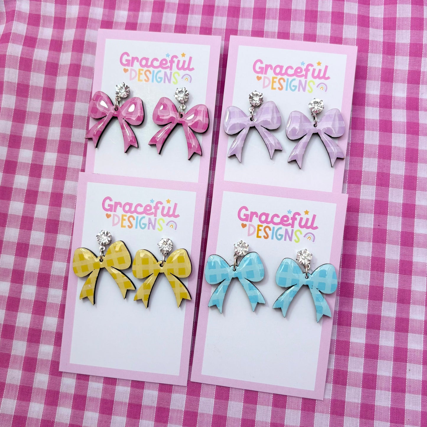 Three pairs of bow-shaped earrings on cards with 'Graceful Designs' branding against a pink checkered background.