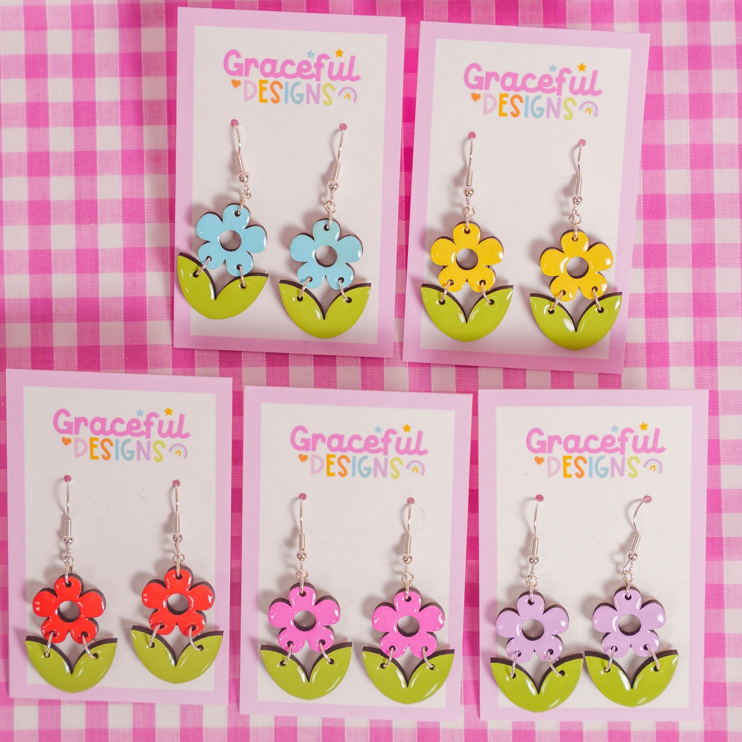 Flower-shaped earrings on display with 'Graceful Designs' packaging against a pink checkered background.