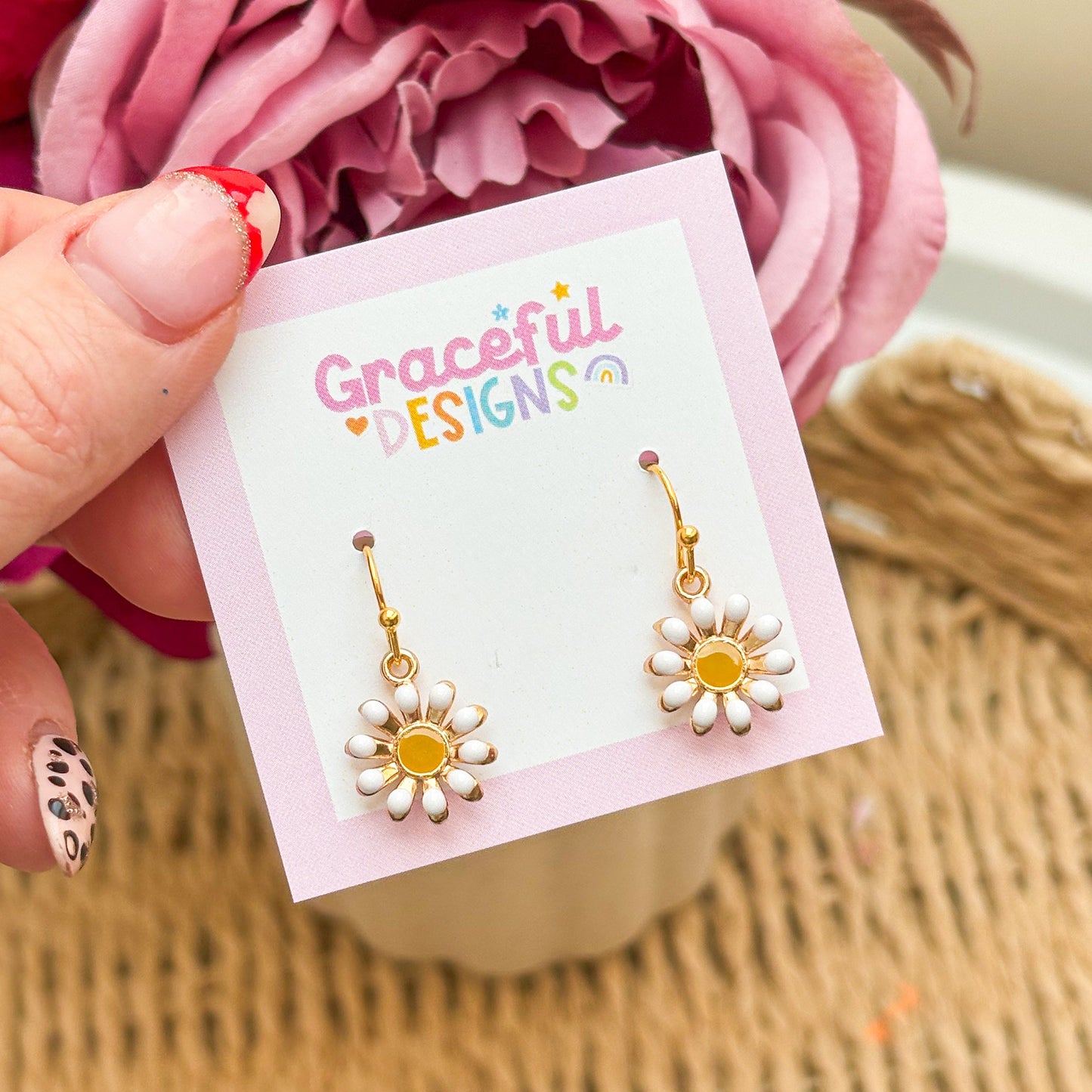 Gold flower-shaped earrings on a 'Graceful Designs' card with pink flowers in the background