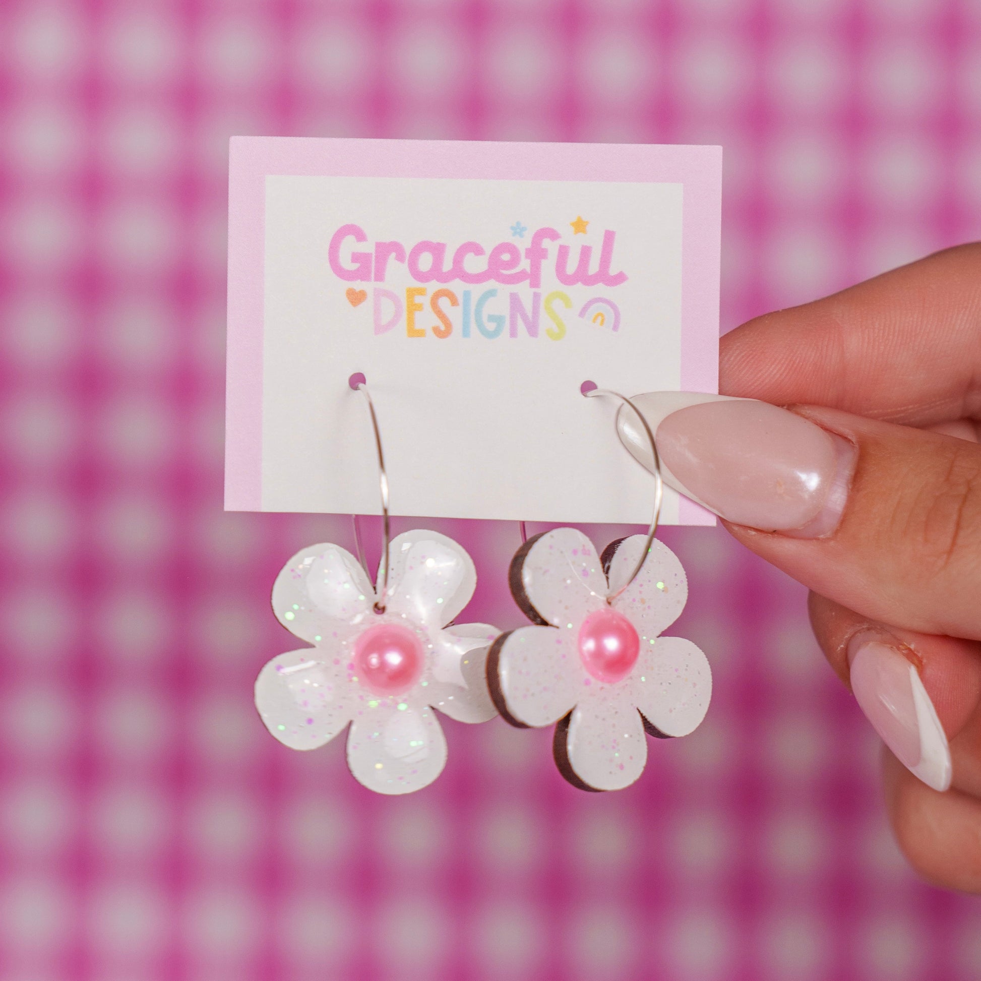 Flower-shaped earrings with pink centers held by a hand against a pink checkered background.