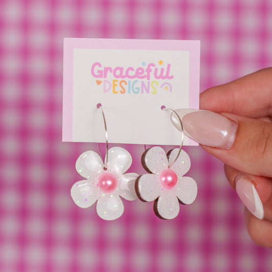 Flower-shaped earrings with pink centers held by a hand against a pink checkered background.