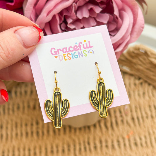 Gold cactus-shaped earrings on a 'Graceful Designs' card with pink flowers in the background.