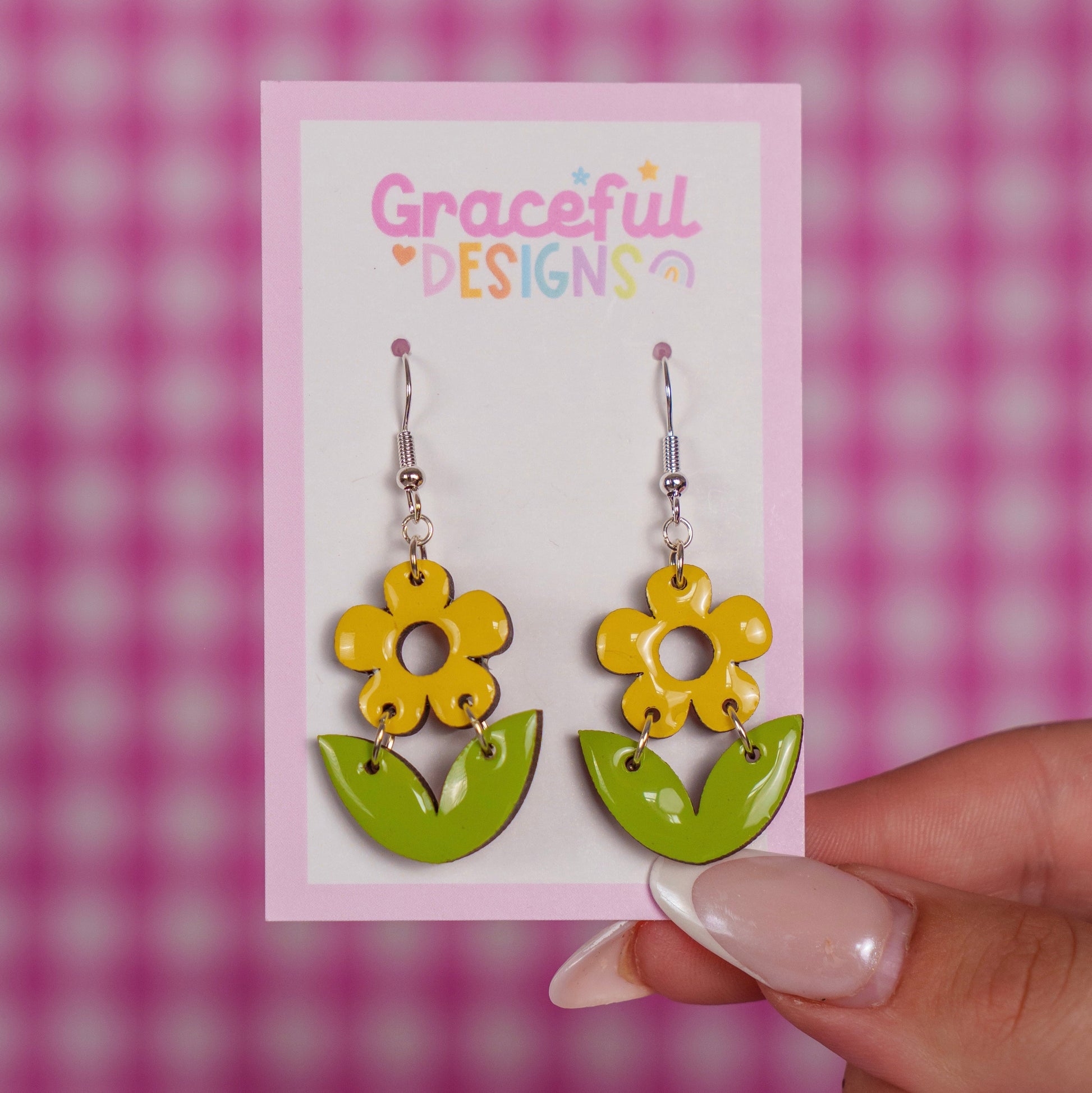 Yellow flower-shaped earrings on a card with 'Graceful Designs' text, held against a pink and white checkered background.