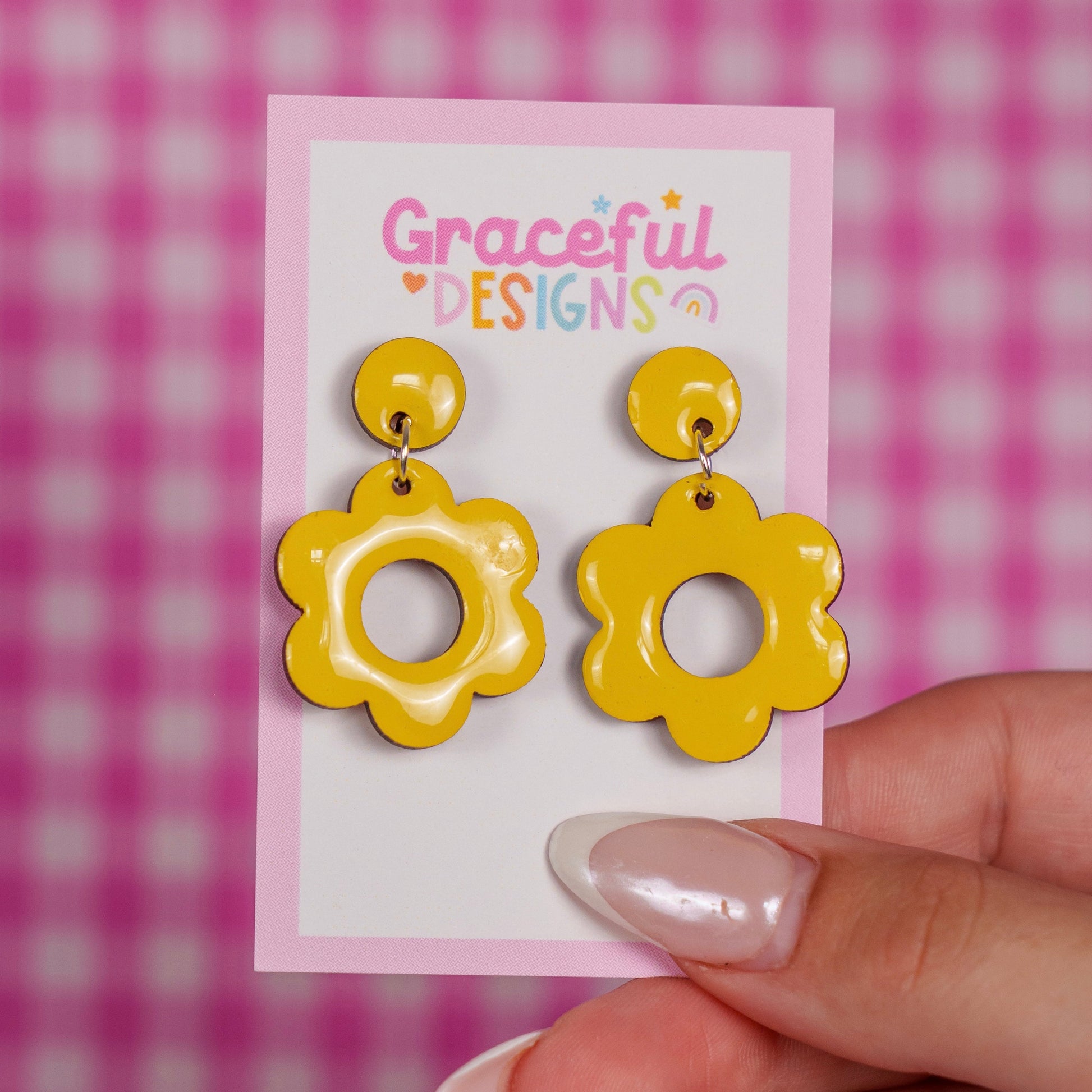 Yellow flower-shaped earrings on a 'Graceful Designs' card against a pink checkered background.