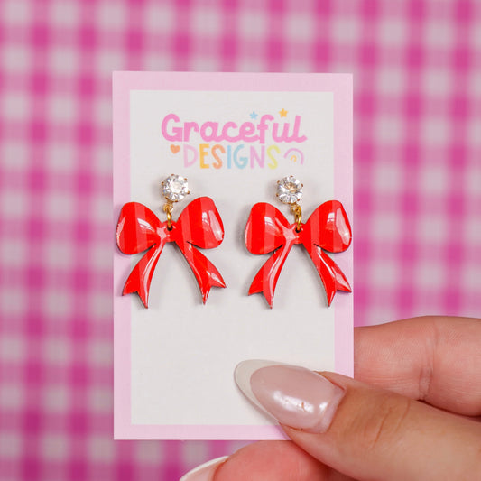 Red bow-shaped earrings on a card held by a hand against a pink checkered background