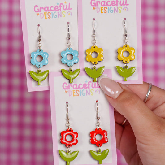 Colorful floral earrings on 'Graceful Designs' cards held by a hand against a pink checkered background.