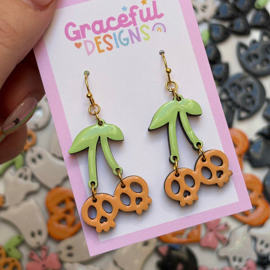 Cherry and skull earrings on a 'Graceful Designs' card with Halloween-themed decorations in the background.