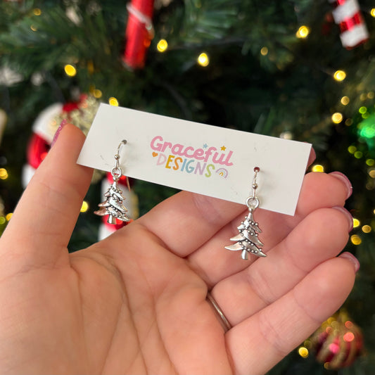 A pair of silver hoop earrings designed to resemble Christmas trees, held in a hand against a festive background with Christmas lights and decorations.