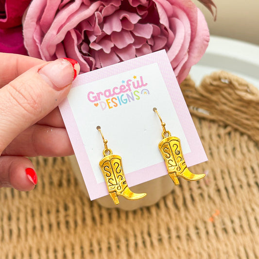 Gold cowboy boot earrings on a 'Graceful Designs' card with pink flowers in the background.
