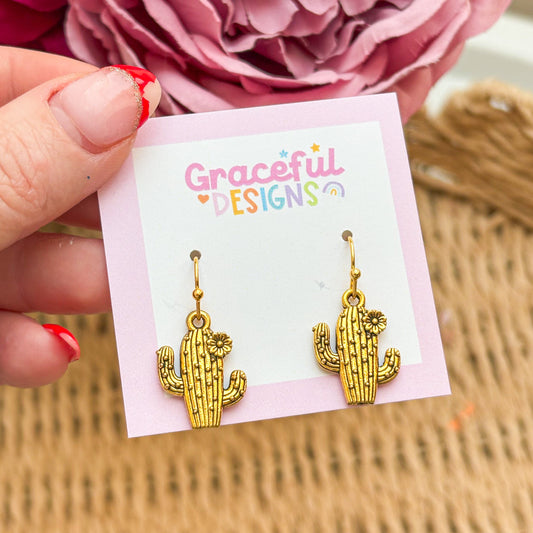 Gold cactus-shaped earrings on a 'Graceful Designs' card with pink flowers in the background.