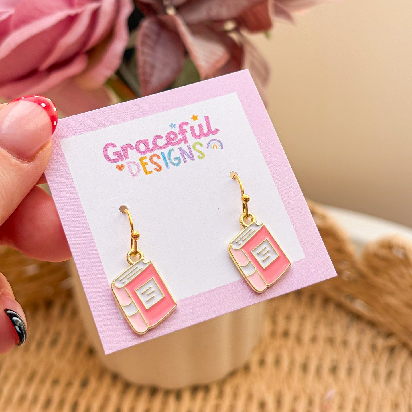 Pink book-shaped earrings on a 'Graceful Designs' card with flowers in the background