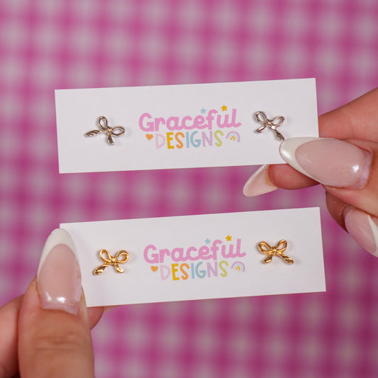 Two pairs of bow-shaped earrings on cards with 'Graceful Designs' branding against a pink checkered background.
