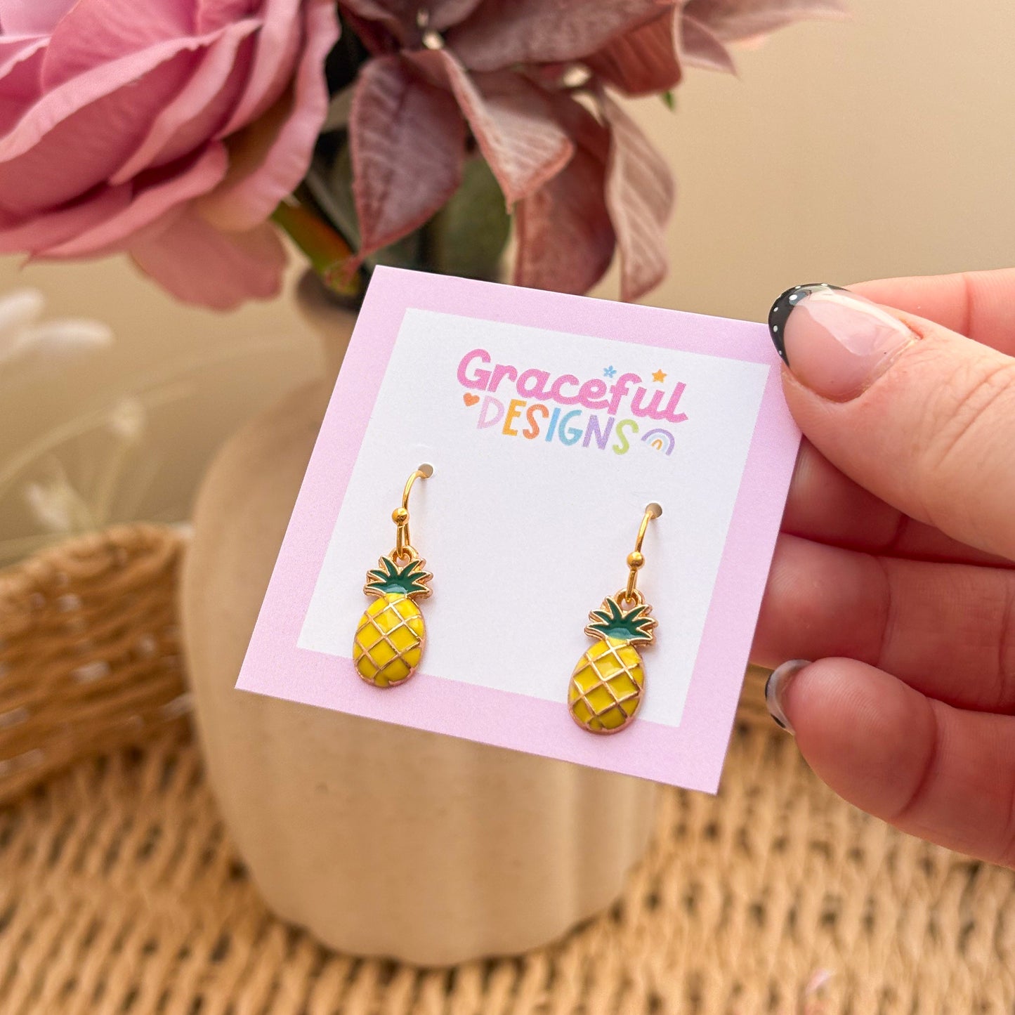 Gold pineapple earrings on a 'Graceful Designs' card with pink flowers in the background