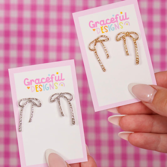 Gold and silver bow-shaped earrings on 'Graceful Designs' cards held by a hand against a pink checkered background.