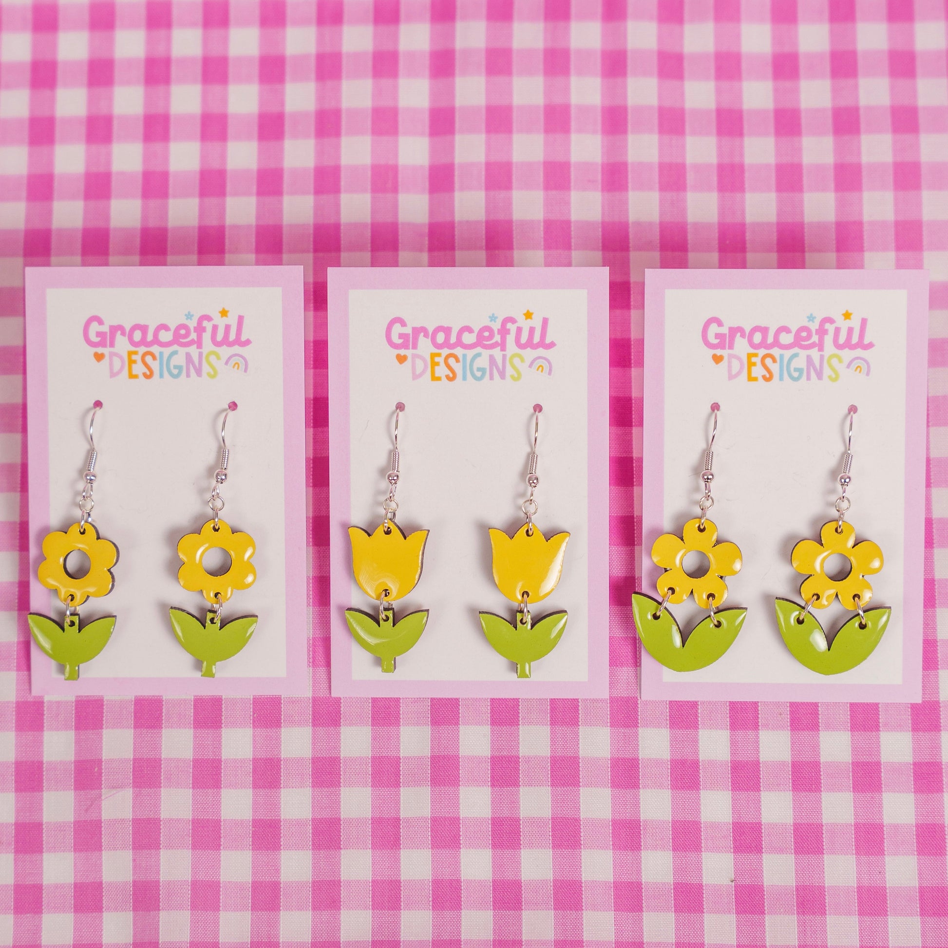 Flower-shaped earrings on cards with 'Graceful Designs' branding against a pink and white checkered background.