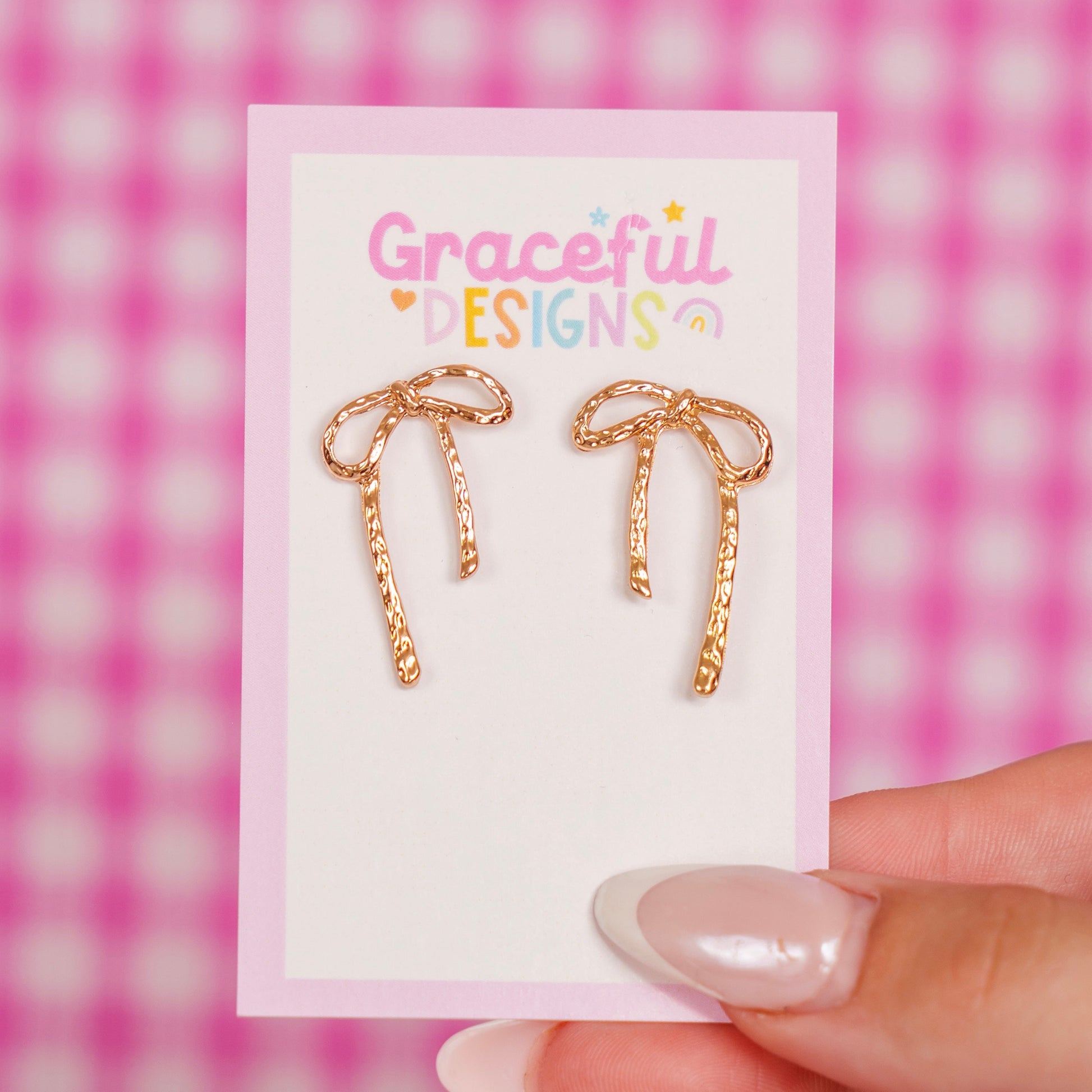 Gold bow-shaped earrings on a 'Graceful Designs' card against a pink checkered background.