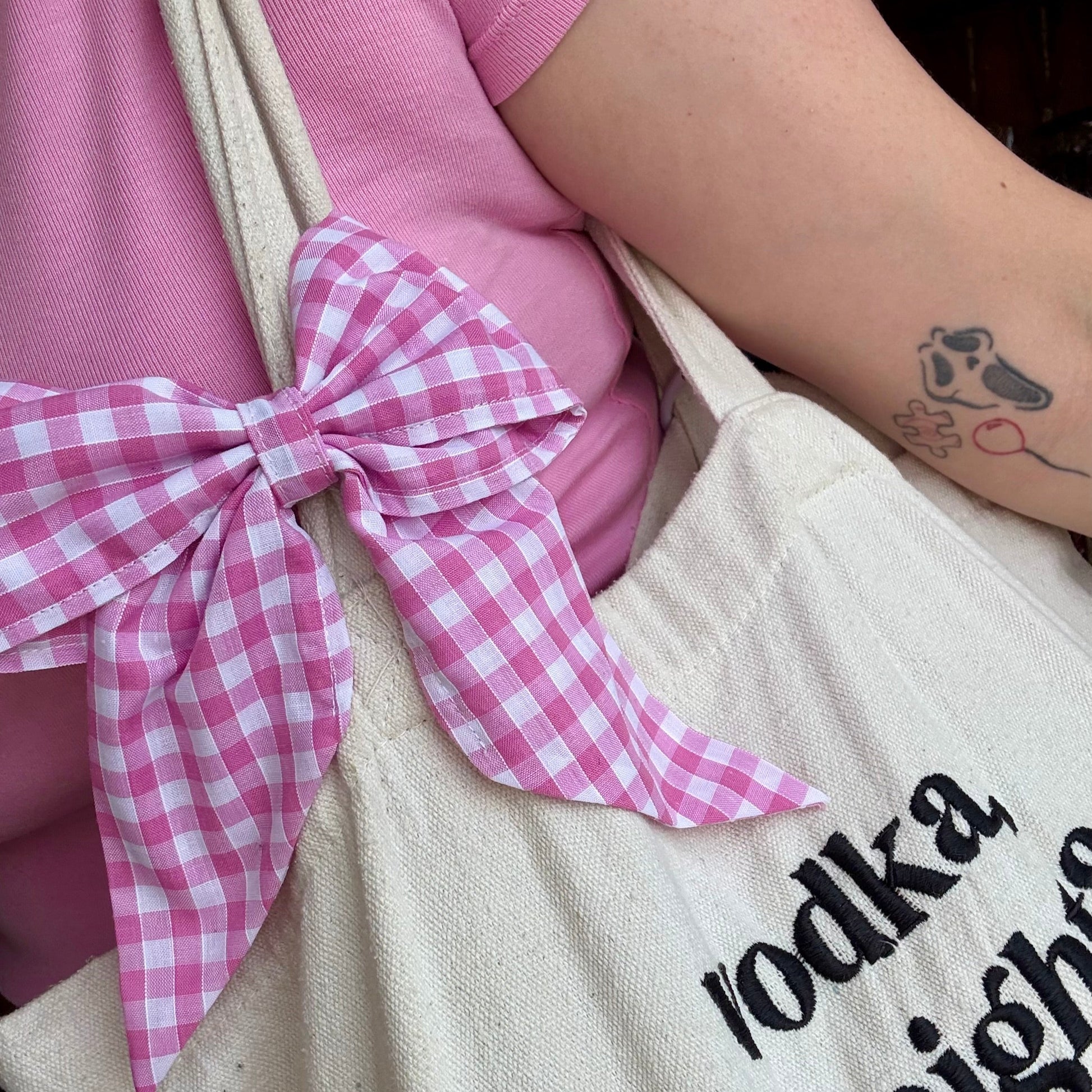 Person wearing a pink shirt with a checkered bow and holding a tote bag with text.