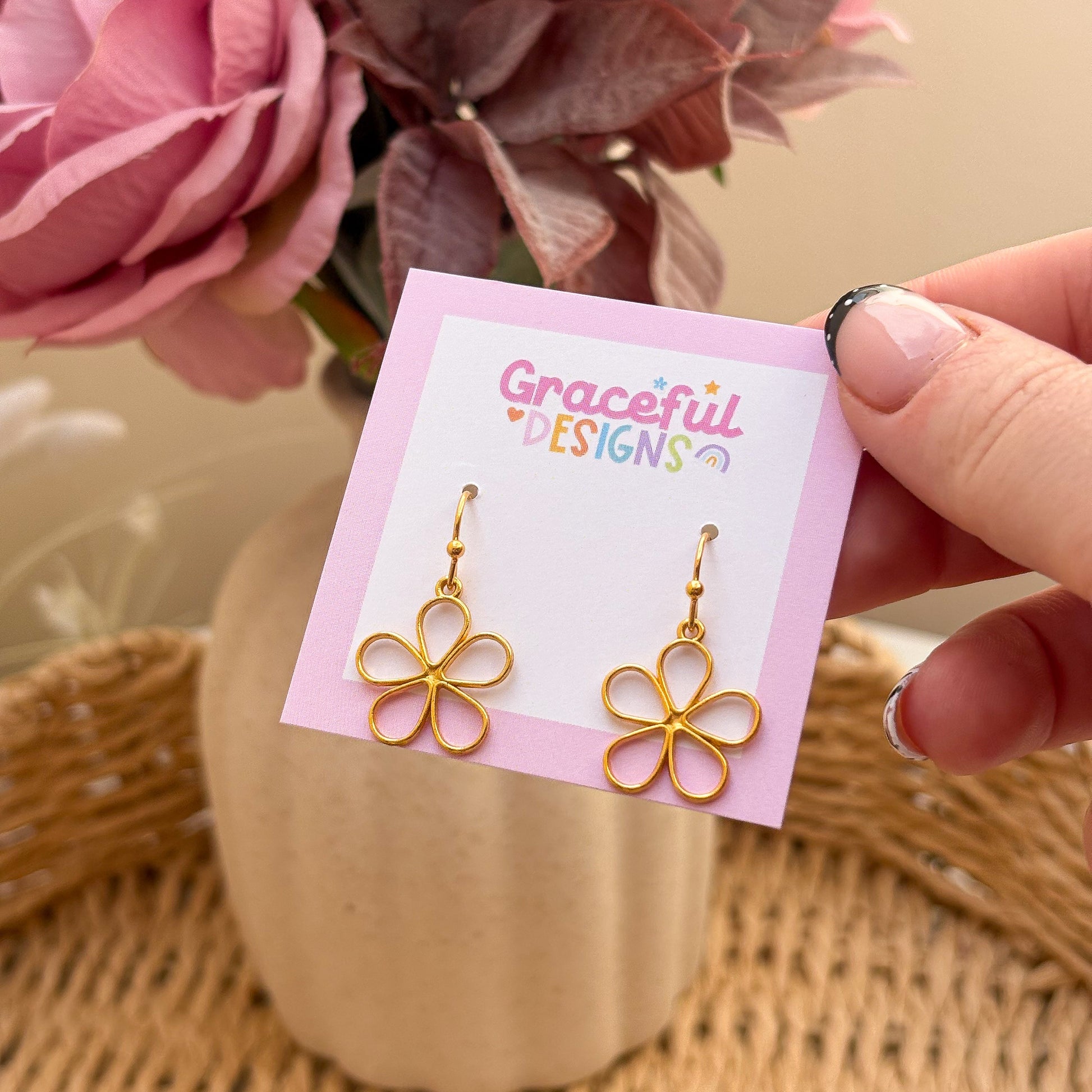 Gold flower-shaped earrings on a 'Graceful Designs' card with pink flowers in the background.