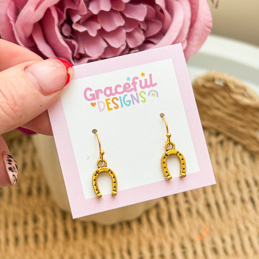 Gold horseshoe earrings on a 'Graceful Designs' card with pink flowers in the background.